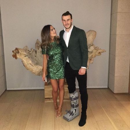 Alba Violet Bale's parents, Gareth Bale and Emma Rhys-Jones are posing for a photo. Emma is wearing green dress and bale is wearing formal caot pant. Bale is smiling at the camera with the injured leg and Emma is looking at him.